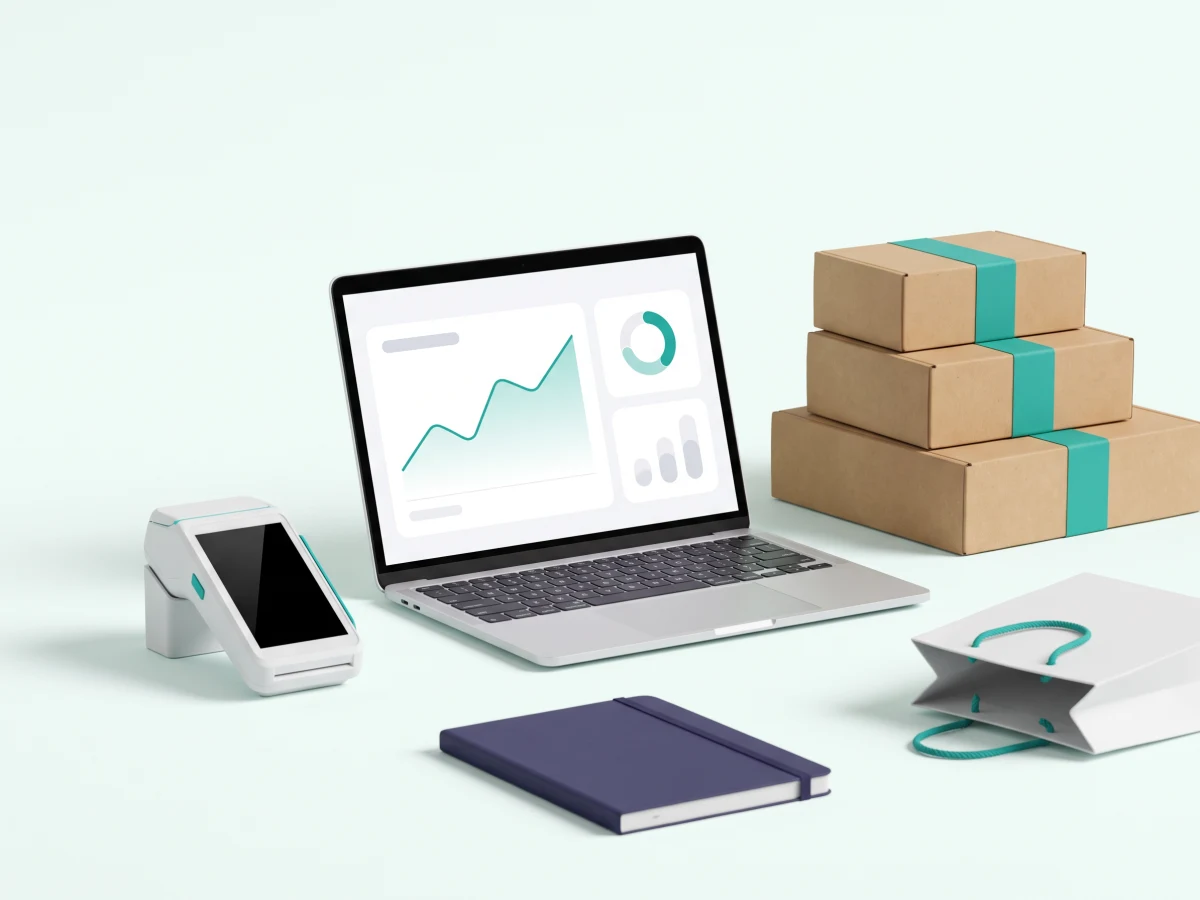 A sales dashboard on a laptop beside a card reader, teal-banded shipping boxes and a shopping bag